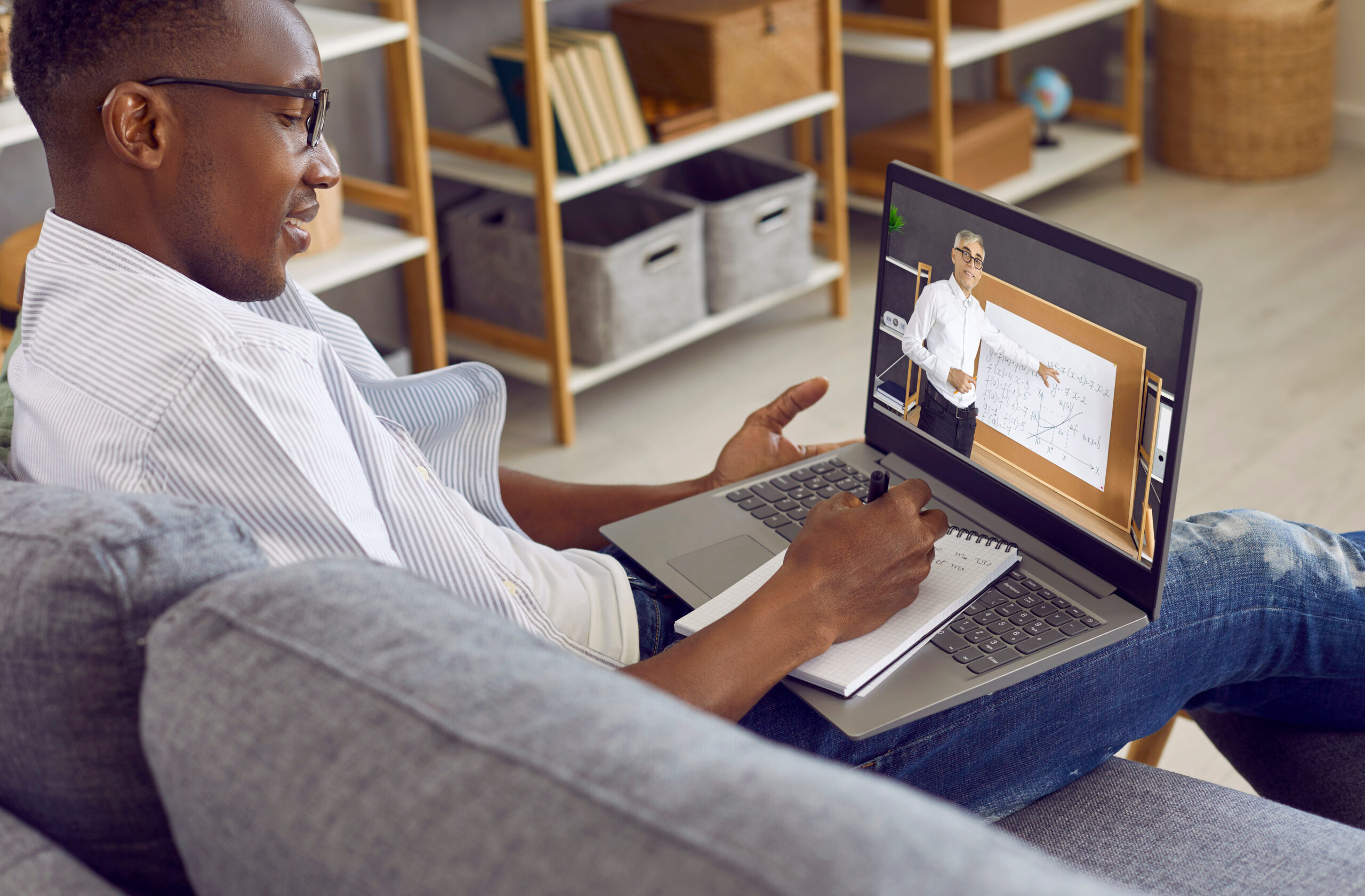 Student having remote class. Happy young African American man sitting on couch at home, using laptop computer, having lesson via video call and taking notes on paper notepad. Online education concept
